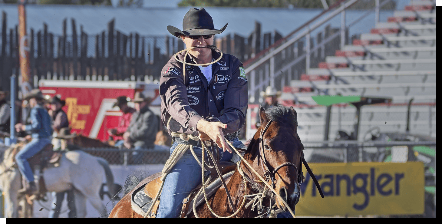 Chet Weitz vistiendo una camisa marrón con parches de patrocinadores y un sombrero negro y gafas de sol, montando a caballo y atando un ternero en un rodeo.